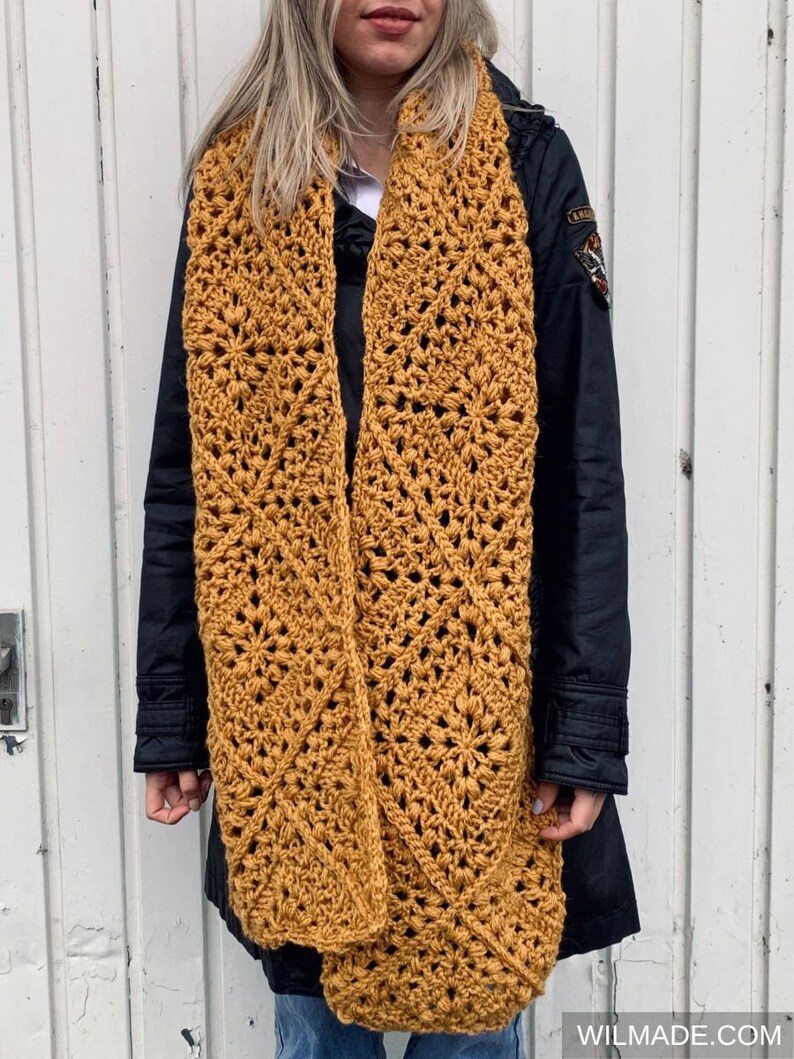 Puede incluir: Una bufanda de crochet de color amarillo mostaza con un patr&oacute;n de diamantes. La bufanda la lleva puesta una persona que lleva una chaqueta negra y unos vaqueros azules.