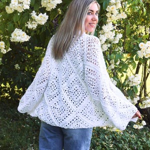 Puede incluir: Un cárdigan blanco de crochet de tejido abierto con mangas largas. El cárdigan presenta un patrón geométrico y se usa sobre unos vaqueros azules. El fondo incluye flores blancas y follaje verde.