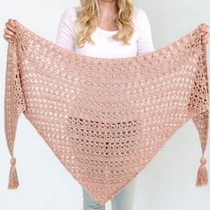 May include: A light peach-colored, crocheted shawl with a triangular shape. The shawl has a delicate open weave design and is held up by a person. The ends of the shawl feature tassels.