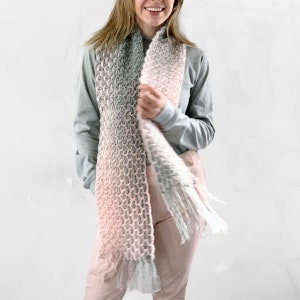 Op de afbeelding: Een zachte, roze en grijze ombre sjaal met franjes. De sjaal is gemaakt van een dik garen en heeft een losse brei textuur.