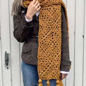 Puede incluir: Una larga bufanda de crochet de color amarillo mostaza con un patr&oacute;n geom&eacute;trico y borlas.