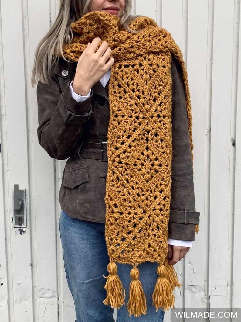 Puede incluir: Una larga bufanda de crochet de color amarillo mostaza con un patr&oacute;n geom&eacute;trico y borlas.