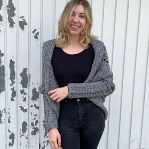 May include: A gray crocheted cardigan with a loose fit and open front. The cardigan has a textured stitch pattern and long sleeves.