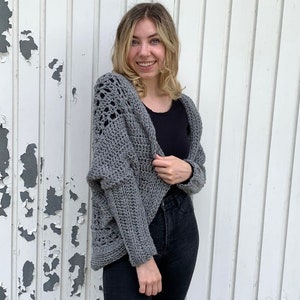 May include: A gray crocheted cardigan with a lacy pattern on the front and sleeves. The cardigan is worn over a black shirt and black jeans.