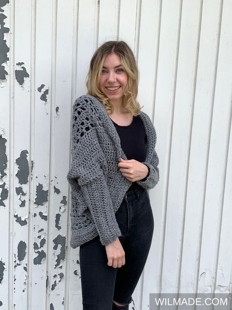May include: A gray crocheted cardigan with a lacy pattern on the front and sleeves. The cardigan is worn over a black shirt and black jeans.