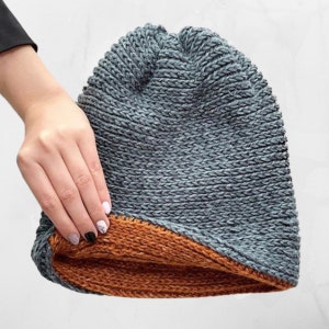 Op de afbeelding: Een grijze gebreide beanie met een bruine fleecevoering. De beanie is omgeslagen om de voering te laten zien.