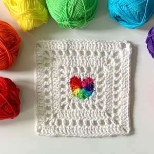 Op de afbeelding: Een wit gehaakt vierkant met een regenboog hartontwerp in het midden. Er omheen liggen bollen garen in verschillende kleuren, waaronder rood, oranje, geel, groen, blauw, paars en roze. Het garen is waarschijnlijk voor handwerk.
