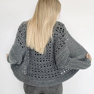 May include: A gray crochet cardigan with a lacy openwork design. The cardigan is shown from the back and has a relaxed fit.