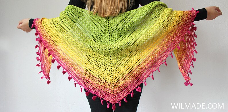May include: A colourful crochet shawl with a gradient of pink, yellow and green. The shawl has a fringe of pink yarn with small heart-shaped pom-poms.