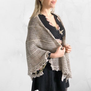 May include: A beige crochet shawl with a scalloped edge. The shawl is draped over a person wearing a black dress with a floral print.