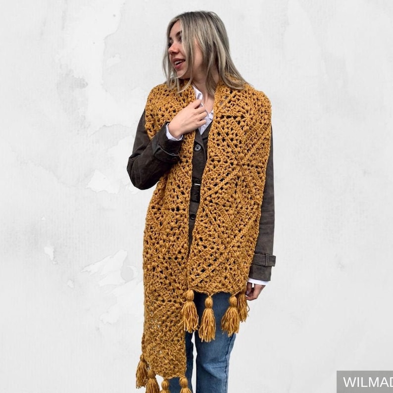 Puede incluir: Una bufanda de crochet de color amarillo mostaza con un patr&oacute;n geom&eacute;trico y borlas. La bufanda la lleva puesta una persona con el pelo rubio y una chaqueta marr&oacute;n.