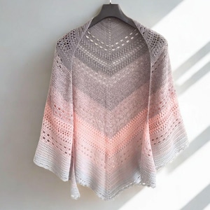May include: A handmade, open-weave shawl in a gradient of colors, from light gray to pink. The shawl is hanging on a hanger against a white wall. The design features a V-neck and a scalloped edge.