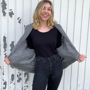 May include: A gray crocheted cardigan with a loose fit and a wide neckline. The cardigan is worn over a black long-sleeved shirt and black jeans.