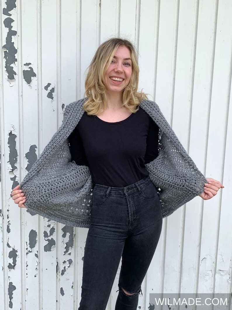 May include: A gray crocheted cardigan with a loose fit and a wide neckline. The cardigan is worn over a black long-sleeved shirt and black jeans.