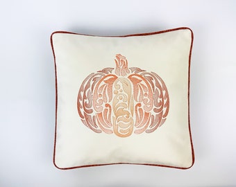 May include: A white throw pillow with a red embroidered pumpkin design. The pumpkin is stylized with a swirling pattern in shades of orange and brown.