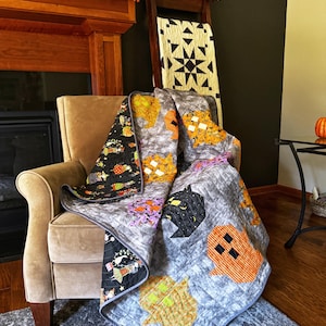 May include: A Halloween-themed quilt draped over a beige armchair. The quilt features a gray background with appliqued orange ghosts, black bats, and other festive designs. A second quilt with a black background and Halloween characters is also visible.