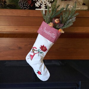 May include: White Christmas stocking with a red and green embroidered design of a girl holding a poinsettia. The stocking has a red felt cuff with the name "Isabella" embroidered in white.