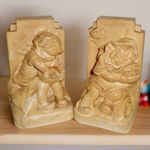May include: A pair of beige bookends, each featuring a sculpted relief of a child holding a bowl. The bookends have a rectangular shape with a decorative top. The overall design has a vintage, antique aesthetic.