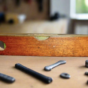 Puede incluir: Un nivel de madera vintage con una parte superior de latón y una ventana de visualización redonda. El nivel es marrón con una parte superior dorada, rodeado de varias herramientas de metal, incluyendo varillas y llaves. Las herramientas están sobre una superficie de madera.