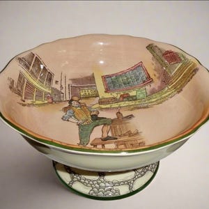 May include: A ceramic bowl with a scalloped edge, depicting a hand-painted scene of a man in a town. The bowl has a pink interior, green and orange trim, and a pedestal base with a decorative pattern.