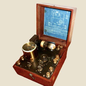May include: A vintage wooden radio device with a hinged lid, featuring a brass cylinder, vacuum tube, and various knobs and switches. The open lid reveals a blue schematic diagram. The device has a dark brown finish.