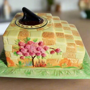 May include: A vintage ceramic butter dish with a yellow and brown checkered pattern. The lid features a black finial and a circular design. The base is a green rectangular plate with a floral design. A pink floral design is on the front.
