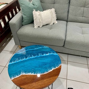 May include: A round wooden table with a blue and white epoxy resin top that resembles ocean waves. The table has black hairpin legs and is sitting on a tiled floor. The letter "D" is written on the floor in front of the table.