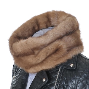 Può includere: Un lussuoso scaldacollo in pelliccia marrone, probabilmente di visone, è drappeggiato attorno al collo di una persona che indossa una giacca di pelle trapuntata nera. La pelliccia è di un ricco colore marrone caldo e la giacca ha dettagli argentati.