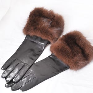 Pode incluir: Um par de luvas de couro preto com punhos de pele castanha. As luvas estão dispostas sobre uma superfície branca, mostrando o seu design elegante e a sua textura luxuosa. Os punhos de pele adicionam um toque de elegância.