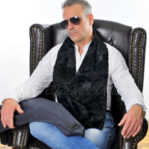 Könnte beinhalten: Ein schwarzer Schal aus Kunstfell mit strukturiertem Muster wird über einem weißen Hemd getragen. Die Person auf dem Bild trägt eine Sonnenbrille und sitzt in einem braunen Ledersessel. Eine dunkelgraue Jacke liegt auf dem Schoß.