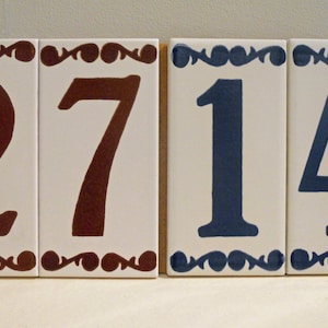 May include: Four white ceramic tiles displaying the numbers 2714. The numbers are in a bold, stylized font. The numbers 2 and 7 are brown, while 1 and 4 are navy blue. Each tile has a decorative border.