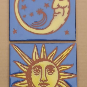 May include: Two ceramic tiles with blue backgrounds. The top tile features a yellow crescent moon with a brown face and stars. The bottom tile features a yellow sun with a brown face and rays.