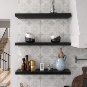 May include: Three matte black floating shelves mounted on a wall with white and gray star-patterned tiles. The shelves hold stacked white and black bowls, wooden pepper mills, a gold cup, a digital thermometer, salt and pepper shakers, and a light blue pitcher.