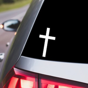 May include: White vinyl decal of a cross on a black background. The decal is applied to the rear window of a silver car.
