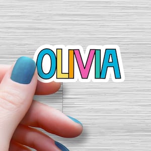 May include: A colorful sticker with the name "OLIVIA" written in bold, outlined letters. The letters are in different colors: blue, yellow, orange, pink, and teal.