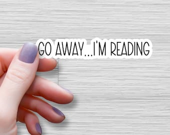 May include: A white sticker with black text that reads "GO AWAY...I'M READING". A hand is holding the sticker.