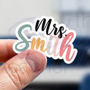 May include: A clear sticker with the text "Mrs. Smith" in colorful, stylized lettering. The sticker has a white border and is held by a hand.