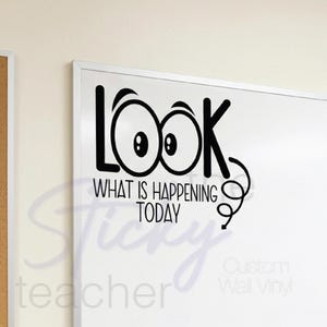 May include: A white dry-erase board with a black vinyl decal that reads "LOOK" with stylized eyes, and "WHAT IS HAPPENING TODAY". The board has a silver frame and a row of colored markers and an eraser.