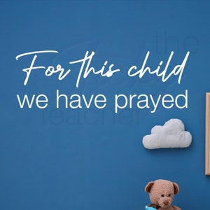 May include: A blue wall with the text "For this child we have prayed" in white. A wooden toy wagon with a teddy bear, a dinosaur, and a pillow inside. A blue chair with a moon-shaped pillow. The wall also has two white clouds.