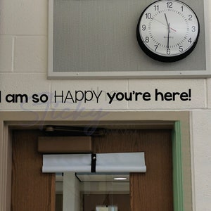 May include: A wall decal with the text "I am so HAPPY you're here!" in black lettering. A round clock with a black frame is mounted above the text. The background is a white brick wall.