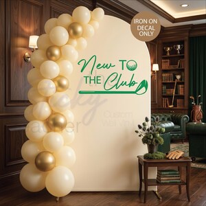 Puede incluir: Un tel&oacute;n de fondo color crema con el texto "New to the Club" en verde, con un dise&ntilde;o de pelota y palo de golf. Un arco de globos crema y dorados enmarca el tel&oacute;n de fondo. Las palabras "Iron on decal only" est&aacute;n en un c&iacute;rculo.