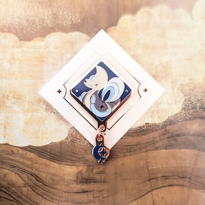 Puede incluir: Un pin de esmalte dorado con una criatura estilizada azul y blanca con una luna creciente en la cabeza. El pin está unido a una cadena dorada con un pequeño amuleto de esmalte azul y blanco.