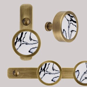 May include: Three brass cabinet knobs with a white marble-like design. The knobs are round and have a raised edge around the marble design. The knobs are shown in different configurations, including a single knob, a knob on a short post, and a pair of knobs on a bar.