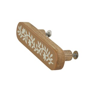 May include: An elongated, rectangular wooden drawer pull with rounded edges. The front face features a light brown finish with a white, intricate floral vine pattern running horizontally. Two metal screws extend from the back for mounting.