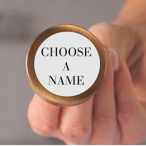 May include: A circular brass wax seal stamp featuring a flat white face with the black text CHOOSE A NAME printed in a serif font. The metallic edge of the stamp has a brushed finish, and the circular design is centered on the stamp face.
