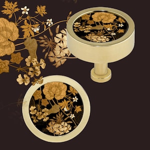 May include: Two gold-toned cabinet knobs with a floral design. The knobs have a circular face with a black background and gold floral pattern. The knobs are set against a dark brown background with gold floral accents.