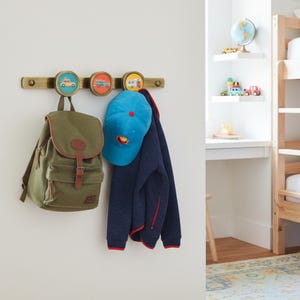 May include: A wall-mounted coat rack with three circular hooks featuring vehicle illustrations. A green backpack, a blue baseball cap, and a navy blue jacket hang from the hooks. The rack is mounted on a white wall.