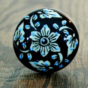 May include: Black ceramic knob with a white floral design and blue accents. The knob features a large white flower with a black center and smaller white flowers and leaves.