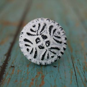 May include: White and black ceramic knob with a decorative floral design. The knob is round and has a raised edge.