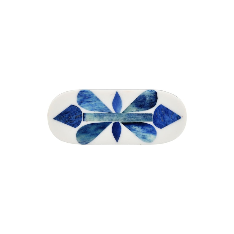 May include: An oval-shaped hair clip featuring a white base with an inlaid pattern of blue and teal geometric shapes. The design consists of a central horizontal bar flanked by four petal-like forms and two pointed diamond shapes on the ends.